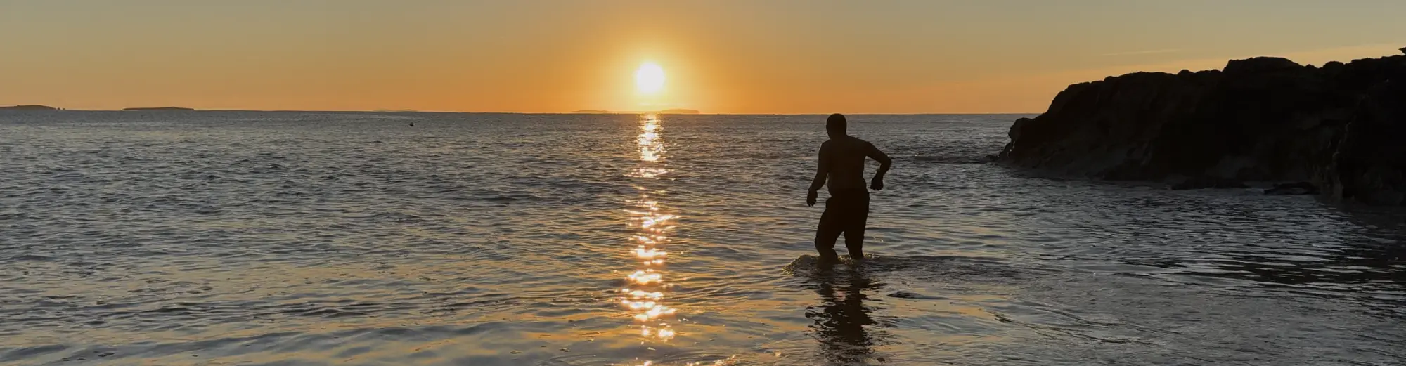 A silhouette of Kenn standing in shallow ocean water during a sunset, with the sun reflecting on the water's surface and rocky formations on the right side.