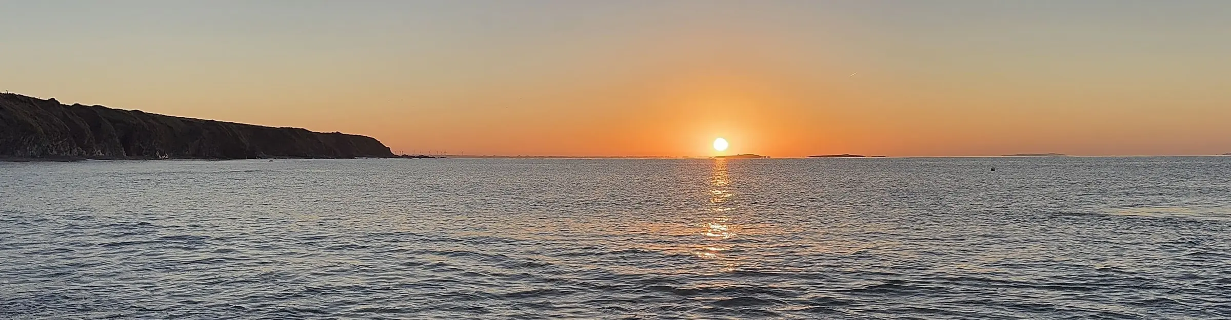 Sunset over the Irish Sea with a clear sky, distant landforms on the horizon, and gentle waves in the foreground.