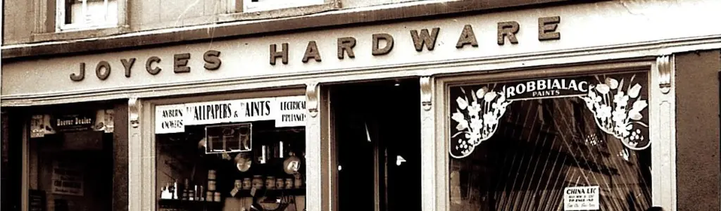 A vintage hardware store facade with the sign "Joyces Hardware." The window displays various tools and paints, including Robbialac paint.