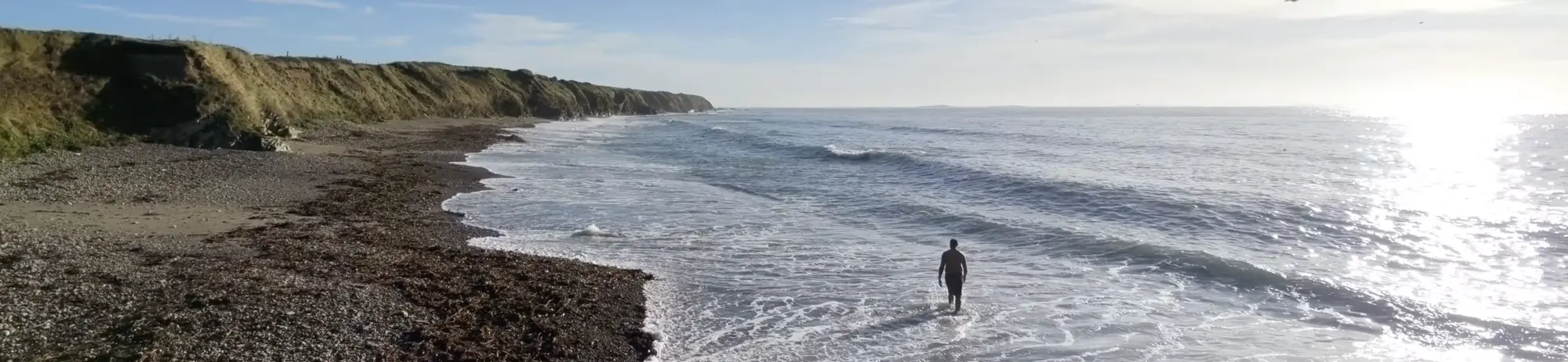 Kenn stands at the edge of the ocean near a rocky shore under a clear blue sky.
