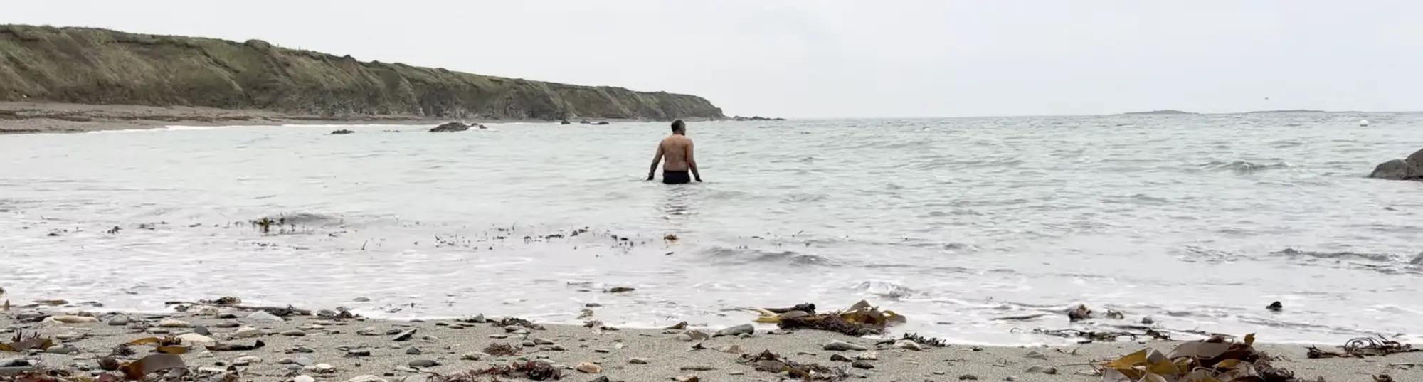 A person wades into the sea from a rocky beach on a cloudy day, with cliffs visible in the distance.