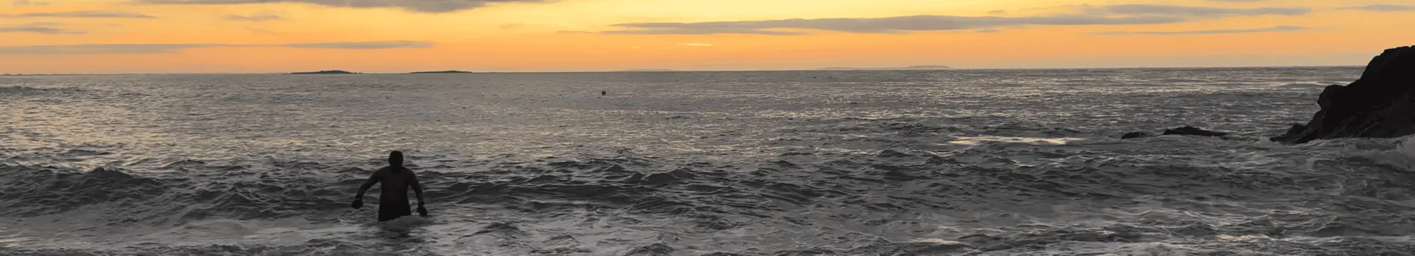 A person stands in the sea at sunset with a rocky shore on the right.