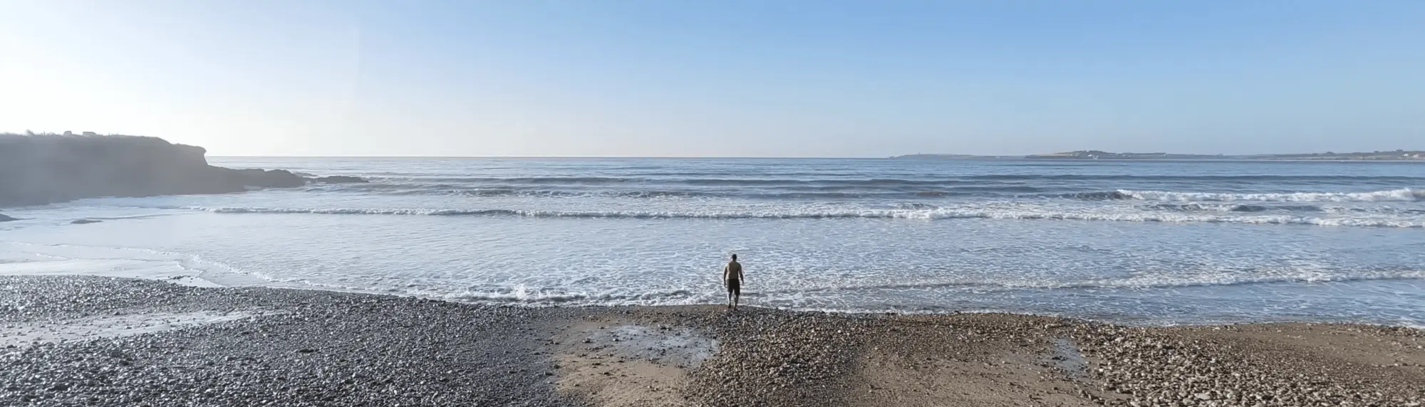 A person stands on a rocky beach facing the ocean waves on a clear day.